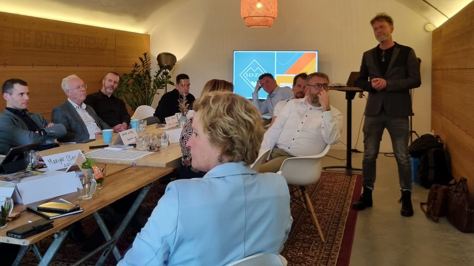 Mensen aan een lange tafel en persoon die Powerpoint-presentatie geeft